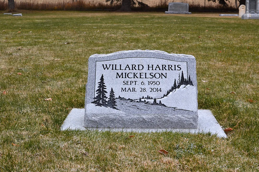 An individual slant marker in gray granite engraved with a mountain and evergreen scene.