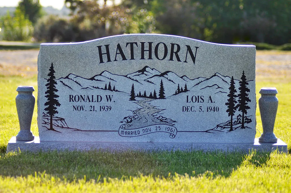 A companion slant marker in gray granite with an engraved mountain scene and a vase at each side.